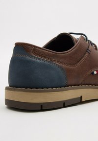 Zapato de cuero marrón con un acento texturizado azul en el talón, suela de color claro en contraste y cordones azul marino, con costuras visibles.
