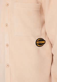 Veste polaire beige à boutons avec poche poitrine et écusson rond noir et orange portant l'inscription "BEYOND" sur le côté droit.