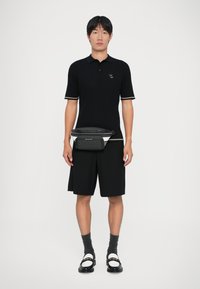 Giovane uomo che indossa un polo a coste nera, pantaloni corti neri, una borsa a tracolla nera e bianca, calzini grigi e mocassini neri con catena dorata su sfondo bianco.