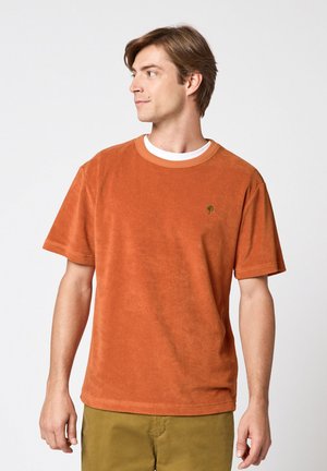 Homme portant une chemise en éponge à manches courtes couleur rouille-orange sur un sous-vêtement blanc et un pantalon vert olive, regardant vers sa gauche.