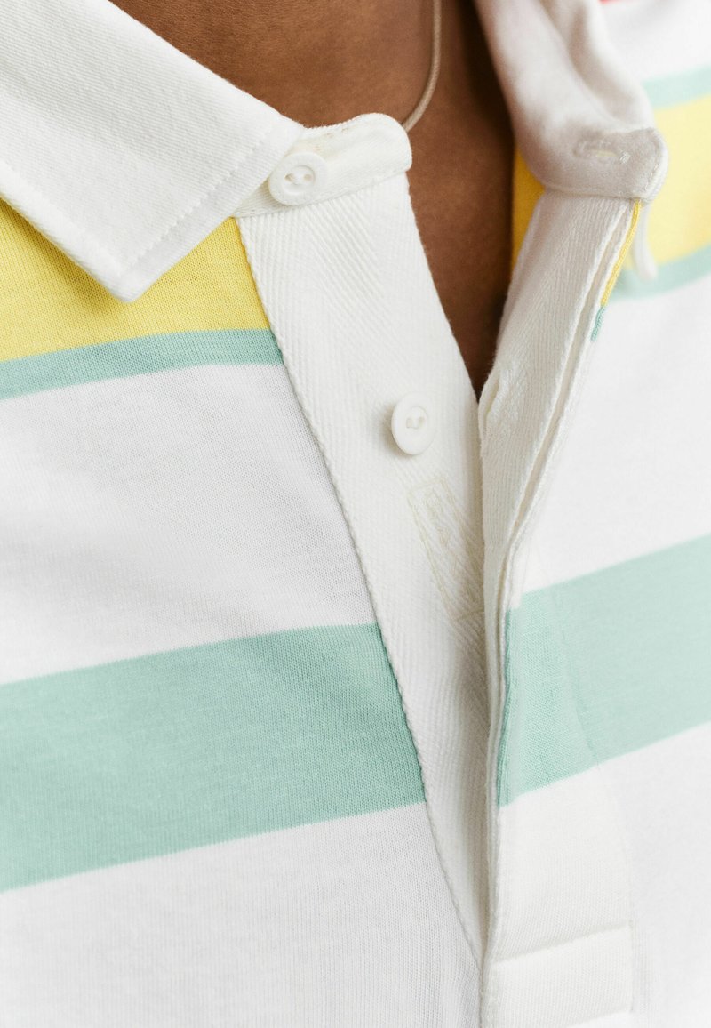 Primo piano di una polo bianca con strisce orizzontali gialle, verdi e azzurro chiaro, chiusa con bottoni bianchi su carnagione marrone.