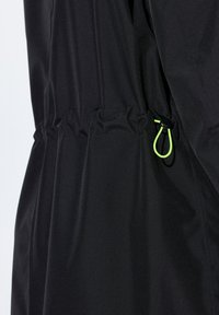 Svart jacka med elastisk dragsko i midjan, med en kontrasterande grön snäpplås. Slät tyg med minimal textur.