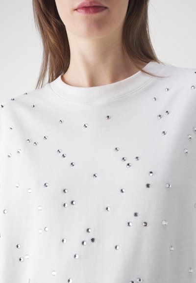 White sweatshirt with a crew neckline, featuring scattered silver rhinestones creating a textured, decorative pattern. Soft fabric.