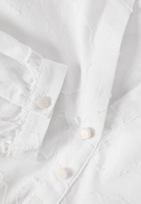 Gros plan sur un tissu blanc avec des motifs de feuilles texturées et trois petits boutons ronds blancs sur un poignet et une patte de chemise.