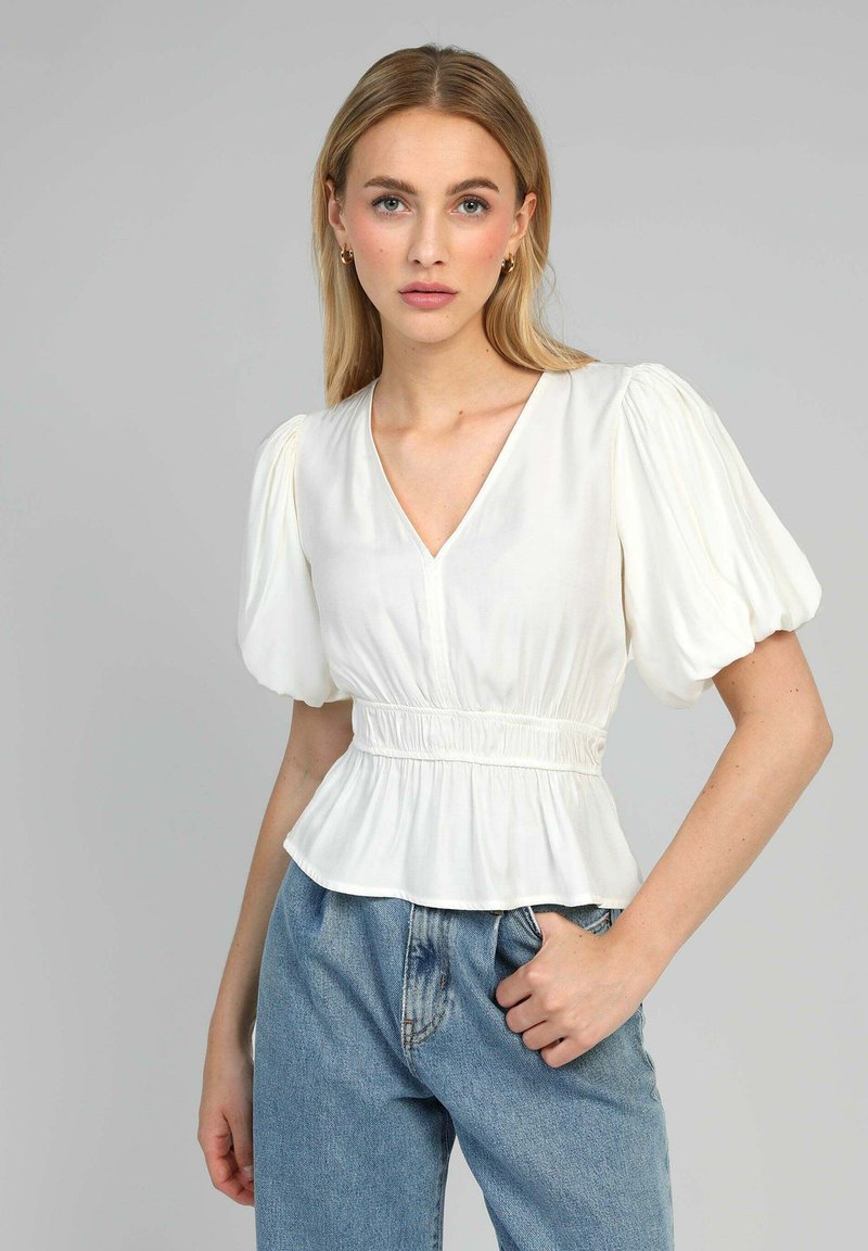 Femme aux cheveux blonds raides portant un blouse blanche à manches bouffantes avec un col en V et une taille froncée, assortie à un jean en denim bleu clair.