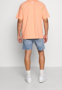 Camiseta melocotón de manga corta con logo blanco en la parte posterior, combinada con pantalones cortos de mezclilla azul y zapatillas blancas.