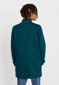 Persona con dreadlocks cortos, vistiendo un abrigo largo color teal y jeans azules, de pie con la espalda hacia la cámara frente a un fondo sencillo.