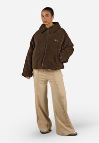 Giacca in pelliccia sintetica marrone con zip e un grande colletto, tasche laterali e un logo discreto. Abbinata a pantaloni beige a gamba larga e calzature grigie.