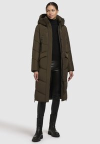 Giacca puffer verde oliva con cappuccio, chiusura a zip davanti e tasche laterali. Presenta un motivo trapuntato e un design lungo fino al ginocchio. Abbinata a pantaloni in pelle neri.