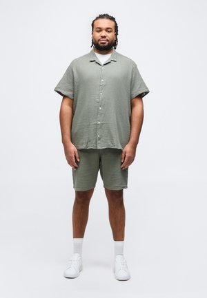 Mężczyzna z zaplecionymi włosami, ubrany w jasnozieloną koszulę na krótki rękaw na guziki oraz dopasowane szorty, białe skarpetki i białe trampki, stojący.