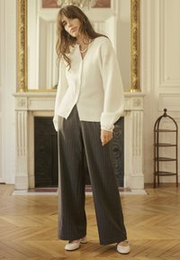 Weißer gestrickter Cardigan mit V-Ausschnitt, der mit Knöpfen geschlossen wird, getragen über grauen, gestreiften weitgeschnittenen Hosen. Das Model trägt cremefarbene Schuhe.