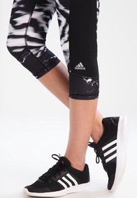 Zwart-wit gemusterde capri leggings met een gladde textuur en een Adidas-logo, gecombineerd met zwarte sneakers met witte strepen.