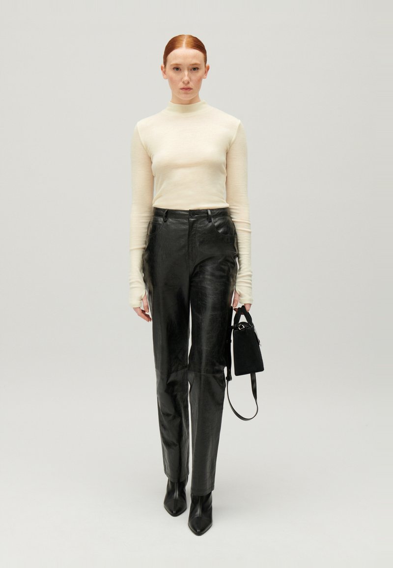 Langarmige crèmekleurige top en zwarte leren broek met hoge taille. Vierkante zwarte laarzen, waarbij een kleine zwarte handtas met een verstelbare schouderband wordt vastgehouden.