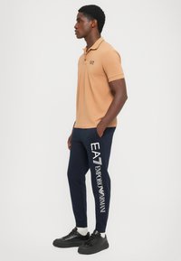 Tan polo shirt with a black logo, paired with navy joggers featuring white “EA7 Emporio Armani” text, complemented by black sneakers.