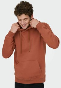 Jonge man glimlacht terwijl hij de capuchon van een roestkleurige hoodie met voorzak aanpast, gekleed in zwarte broek tegen een effen achtergrond.