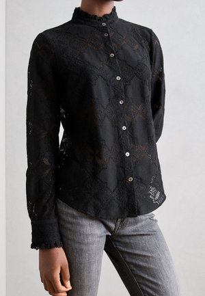 Henkilö pukeutunut mustaan pitsiin, jossa on pitkähihaiset napitettava paita ja korkea kaulus, yhdistetty vaaleanharmaisiin farkkuihin yksiväristä taustaa vasten.