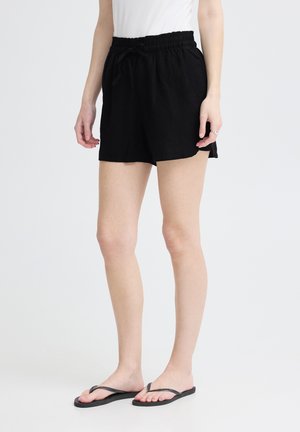 Žena nosiace čierne elastické šortky s šnúrkou v páse a čierne žabky, stojaca na bielom pozadí.