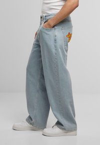 Helle blaue weit geschnittene Jeans aus Denim, mit einer Gesäßtasche, die ein buntes Stickdesign aufweist. Getragen mit weißen Turnschuhen.