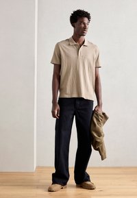 Polo beige a maniche corte, pantaloni neri a gamba larga e scarpe color sabbia. La maglietta presenta una texture sottile e un colletto a due bottoni.