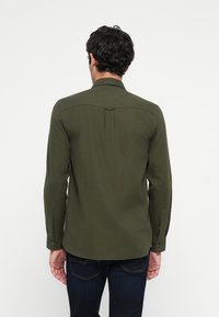 Hombre con cabello rizado oscuro, vistiendo una camisa de manga larga verde oliva y jeans azul oscuro, de pie de espaldas contra un fondo blanco.