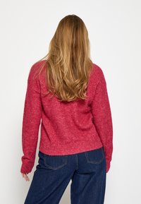 Mikasa ar mīkstu, sarkanu audumu, ar nolaižamām plecu šuvēm, ribotām piedurknēm un apakšmalu, saskaņota ar tumši ziliem, platām kājām džinsiem. Tekstūra izskatās mīksta un silta.