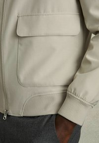 Jachetă bej deschis cu o textură netedă, având un buzunar pe piept, manșete cu nervuri și un fermoar frontal. Designul este casual și elegant.