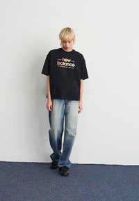 Asmuo su trumpais šviesiais plaukais stovi priešais baltą sieną, vilkėdamas juodą New Balance marškinėlį, išblukusius mėlynus džinsus ir juodus batus, žiūrėdamas žemyn.