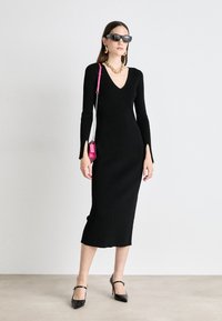 Black ribbed knit dress with long sleeves and side slits. Paired with black heels, sunglasses, and a small pink bag, featuring gold jewelry accents.