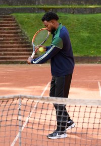 Jogador de ténis com uma jaqueta azul e verde com fecho éclair, calças pretas e sapatos pretos, a preparar-se para acertar numa bola de ténis amarela com uma raquete.