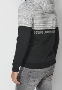 Maglione con cappuccio caratterizzato da una parte superiore grigia testurizzata e una sezione inferiore nera tinta unita. Presenta il testo bianco "KRSHI URBANCON" sul retro. Materiale in cotone misto.
