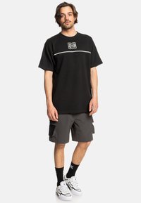 Black short-sleeve t-shirt with white horizontal stripe and logo, paired with dark gray cargo shorts, black socks, and white sneakers.