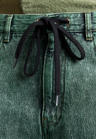 Πράσινες τζιν βερμούδες με μαύρο κορδόνι και ασημένιες λαβές, υφασμάτινη υφή και εμφανή ραφή γύρω από τη μέση και τις τσέπες.