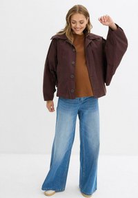 Giacca oversize color bordeaux con ampi revers, bottoni frontali e maniche voluminose, abbinata a un top marrone e jeans a gamba larga blu.