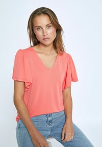 Korálová blúzka s výstrihom do V a krátkymi volánovými rukávmi, vyrobená z ľahkej látky. Modelka má na sebe svetlomodré džínsy a sedí na stoličke na neutrálnom pozadí.