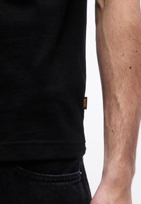 Κοντινό πλάνο του χεριού ενός ατόμου δίπλα σε μαύρο πουκάμισο με μια μικρή πορτοκαλί ετικέτα "BOSS" στο κάτω μέρος, σε συνδυασμό με μαύρο τζιν.