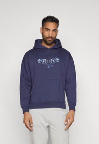 Sudadera azul marino hecha de una mezcla de algodón, con un bolsillo delantero y un estampado gráfico. Con puños y dobladillo acanalados. Llevada sobre pantalones de chándal grises claros.