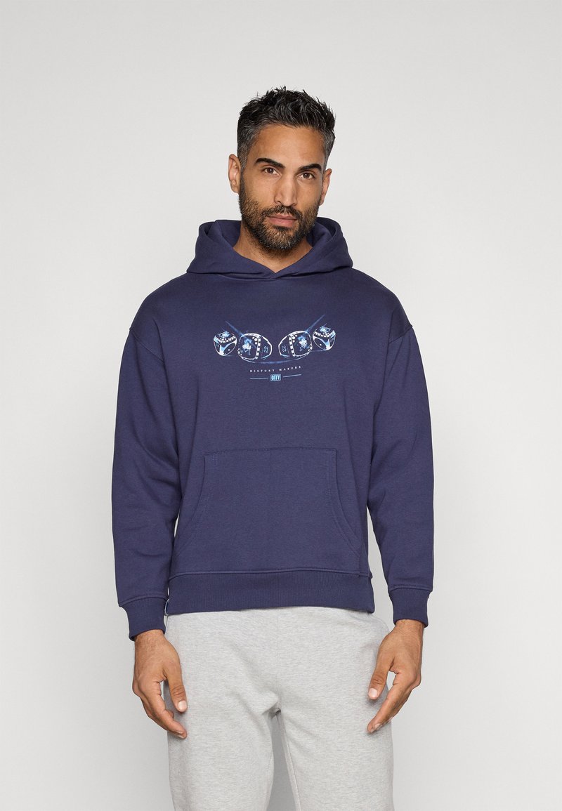 Sudadera azul marino hecha de una mezcla de algodón, con un bolsillo delantero y un estampado gráfico. Con puños y dobladillo acanalados. Llevada sobre pantalones de chándal grises claros.