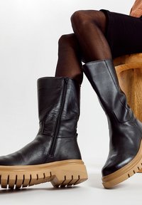 Schwarze Lederstiefel bis zum Knie mit einem Seitenreißverschluss und beiger, dicker Sohle, ausgestattet mit strukturierten Details und einer glatten Oberfläche.