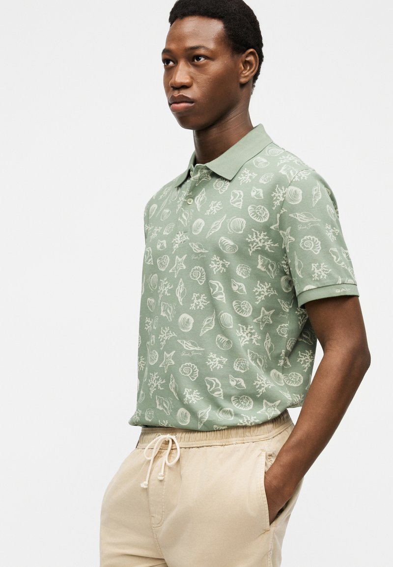 Uomo in piedi con le mani in tasca, indossa una polo verde decorata con conchiglie bianche e pantaloni beige con coulisse.