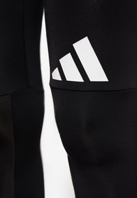 Czarne legginsy sportowe wykonane z elastycznego materiału, z wyraźnym białym logo w postaci trzech pasków na górnej części nogawki.