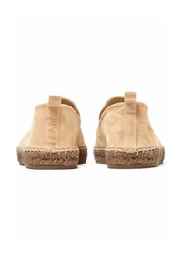 Chaussures slip-on en daim beige de forme arrondie, avec des semelles en jute tressé et de petites languettes au talon. Texture lisse sans embellissements visibles.