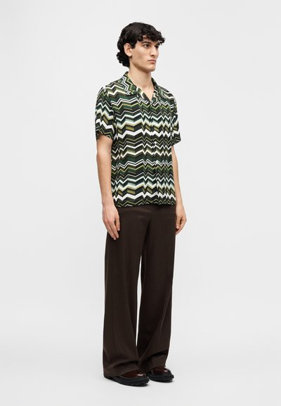 Πουκάμισο με κοντά μανίκια σε σχέδιο ζιγκ-ζαγκ σε πράσινο, μαύρο και άσπρο, συνδυασμένο με καφέ παντελόνι φαρδιάς γραμμής και μαύρα παπούτσια.