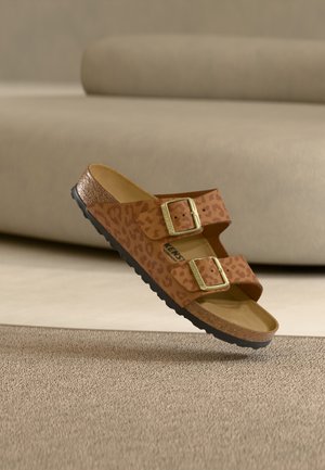 Rudos sandalai su leopardų rašto spauda, dvi reguliuojamos auksinės sagtelės, kamštinė pado dalis ir tekstūruotas juodas guminis padažas, pakelti virš smėlio spalvos paviršiaus.