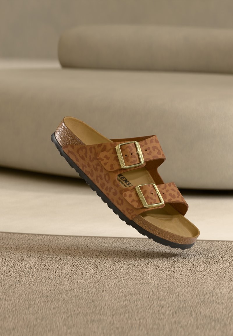 Καφέ σανδάλια με λεοπάρ σχέδιο, δύο ρυθμιζόμενες χρυσές αγκράφες, πάτος από φελλό και υφή μαύρης καουτσούκ σόλας, ανυψωμένα πάνω από μία μπεζ επιφάνεια.