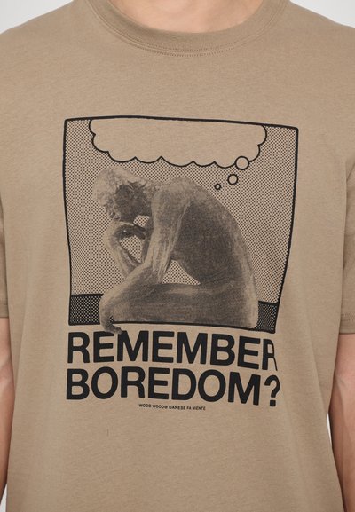 T-shirt en coton beige présentant un graphisme sombre d'une figure assise en pleine réflexion, avec le texte "RAPPELEZ-VOUS DE L'ENNUI ?" en lettres noires grasses en dessous.
