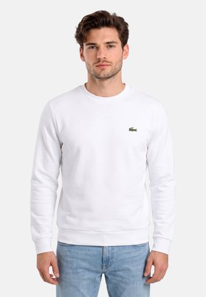 Giovane uomo che indossa una maglietta bianca semplice a maniche lunghe con un piccolo logo di coccodrillo verde e jeans azzurri chiari, in piedi davanti a uno sfondo bianco.