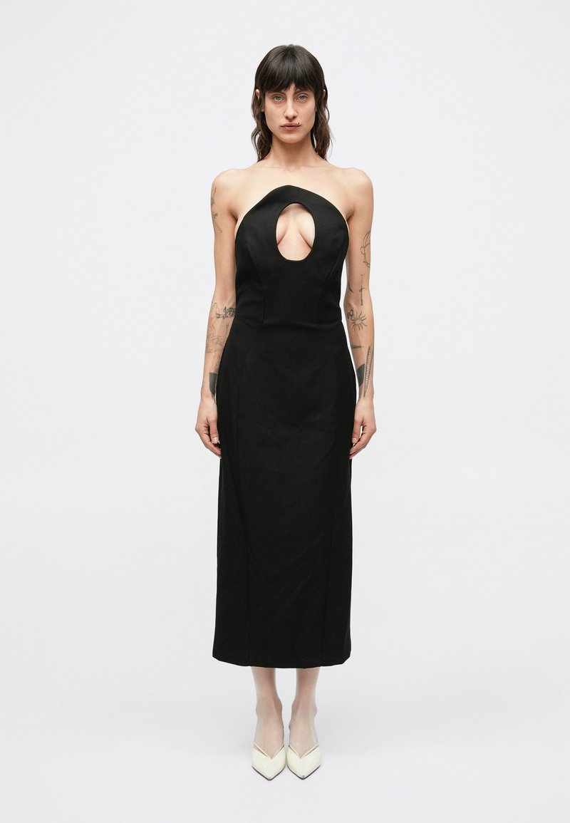 Μαύρο φόρεμα χωρίς τιράντες με βαθύ άνοιγμα μπρο�στά, εφαρμοστή σιλουέτα και μεσαίο μήκος; σε συνδυασμό με λευκές μυτερές γόβες.