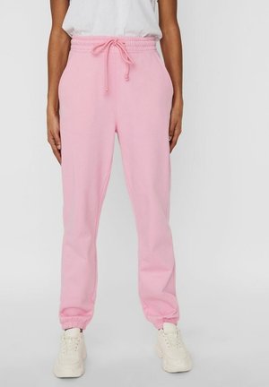 Personne portant un pantalon de survêtement rose clair avec cordon et des baskets blanches, debout devant un fond clair uni.