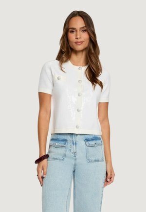 Donna che indossa un cardigan bianco a maniche corte con bottoni e dettaglio con tasca frontale, abbinato a jeans azzurri a vita alta con grandi tasche frontali.