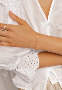 Złota bransoletka z małymi białymi perłami, noszona na opalonej ręce, w zestawieniu z białą wyszywaną koszulą z detalami w kształcie oczek.